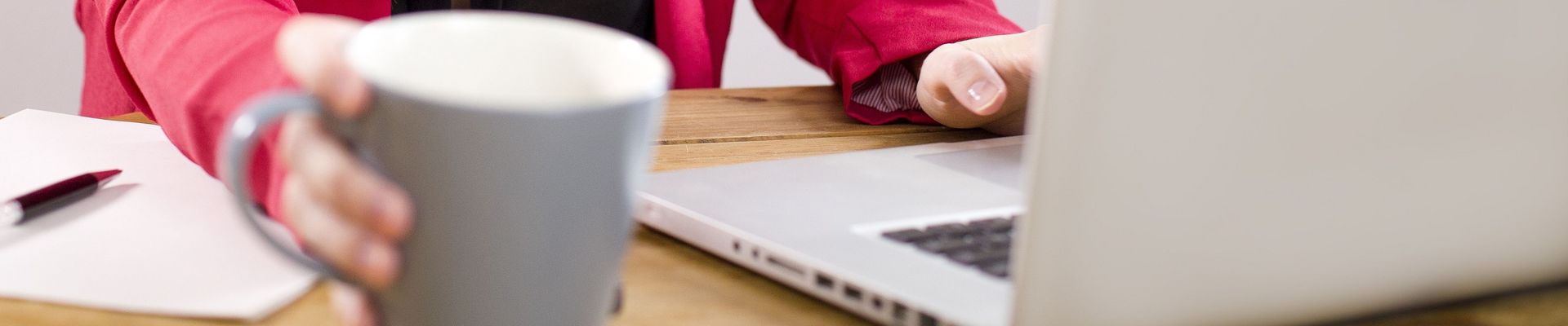 Eine Frau sitzt mit einem Kaffe am Schreibtisch und arbeitet an ihrem Laptop.